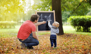 A man and a child outdoors in an autumnal scene, with a blackboard in front of them and the numbers 1 + 1 written in chalk on it. Both man and child are looking at the chalkboard